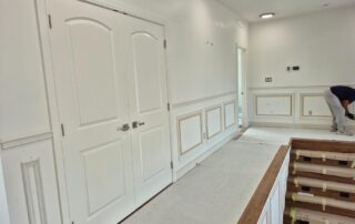 A hallway undergoing interior painting, showcasing new white wainscoting, doors, and protected stairs during renovation.