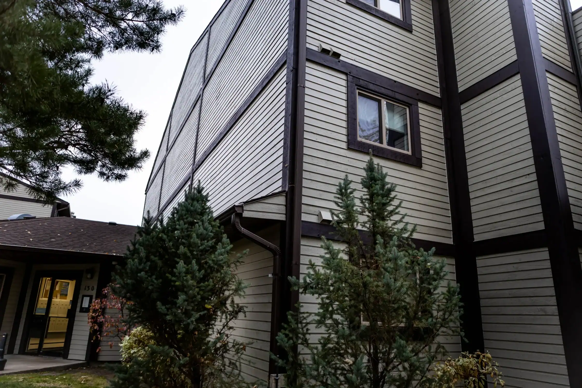 Detailed Exterior Painting for Apartment Complexes Close up of an apartment building corner with light gray siding, dark trim, and evergreen landscaping.