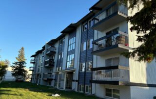 A large apartment building with blue and white siding and balconies, viewed from a sunny, grassy area.