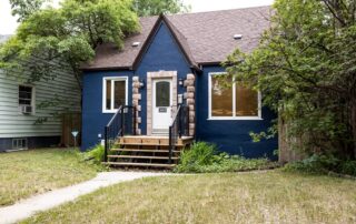 Wide shot of a charming blue stucco house with stone accents, a front porch, and green landscaping.