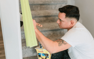 Painter in white shirt applying green painter's tape to white interior wall trim during residential painting prep
