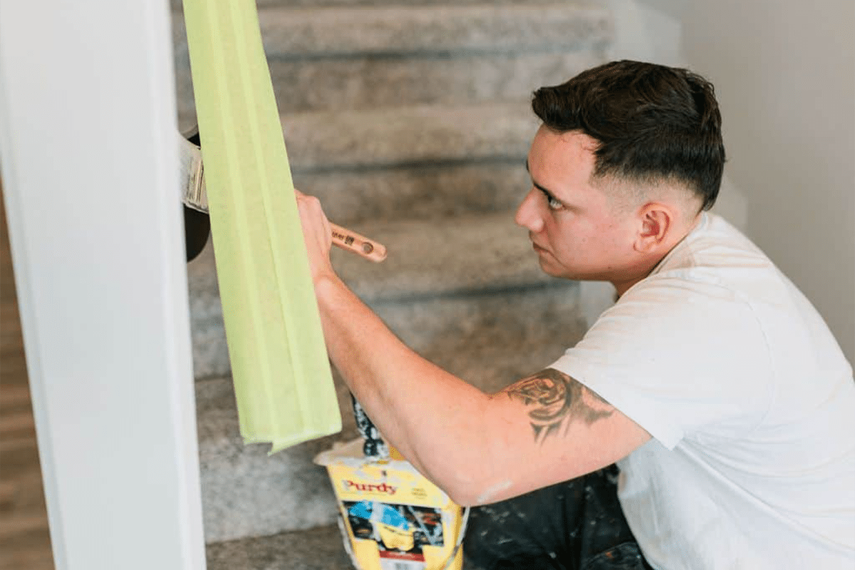 Painter in white shirt applying green painter's tape to white interior wall trim during residential painting prep