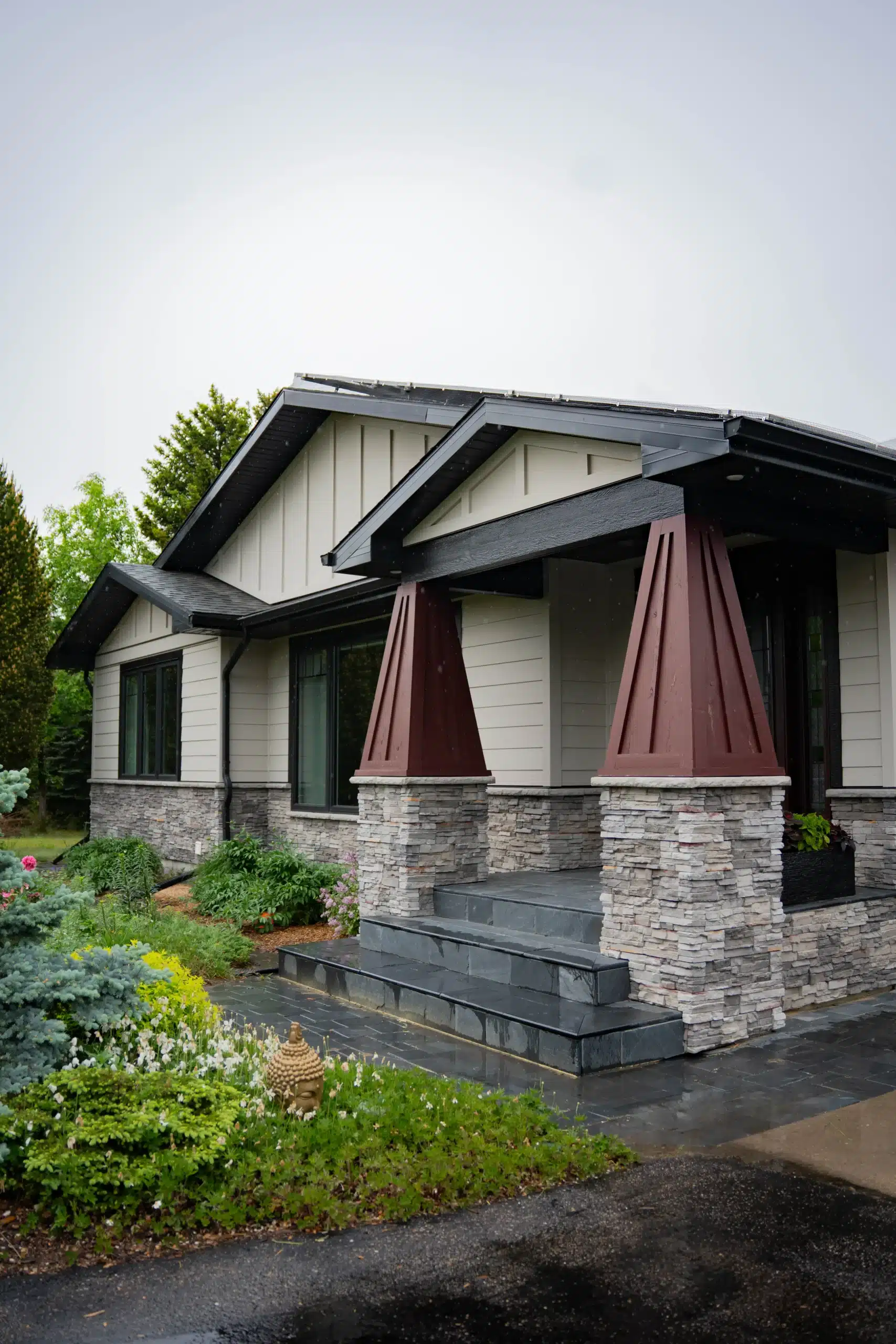 Modern House Exterior with Light Siding Dark Trim and Stone Accents A modern residential house exterior features light beige siding, dark trim, stone pillars, and a landscaped garden.
