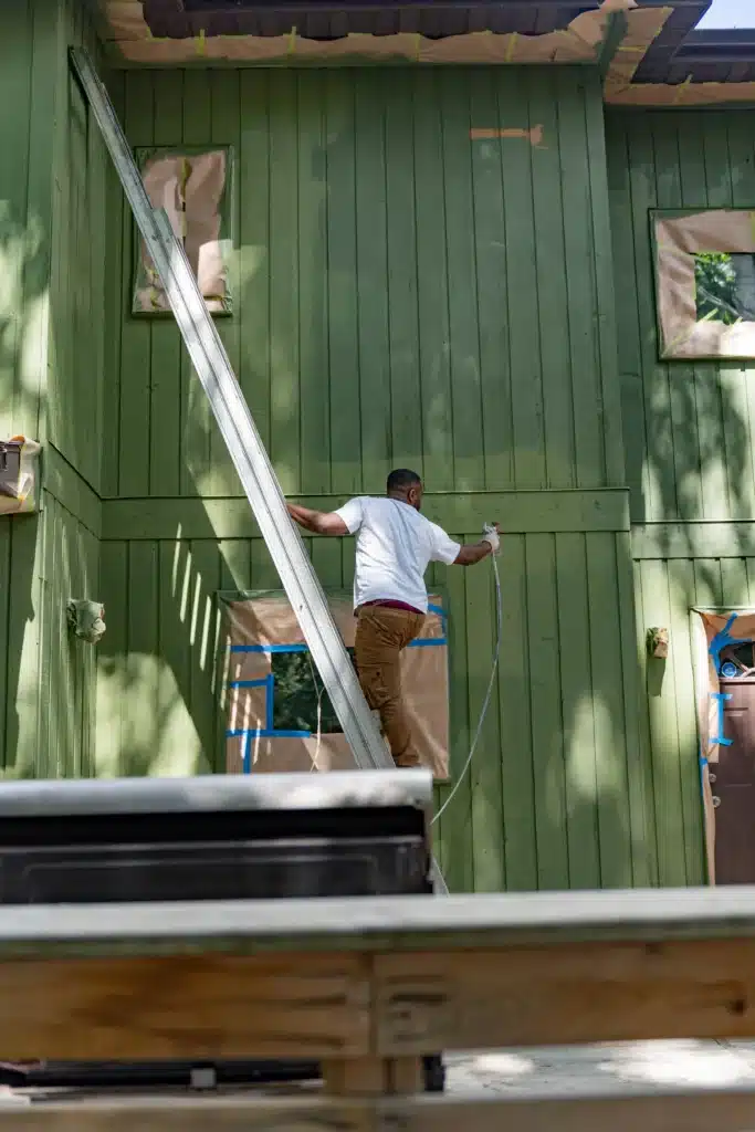 Professional Exterior House Painting Services A professional painter on a ladder spray painting the green exterior of a residential house.