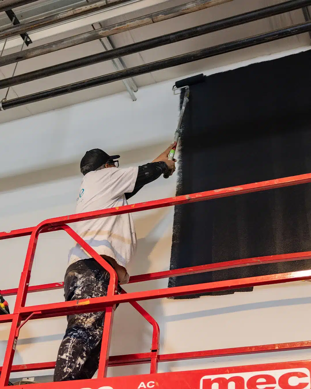 Black Accent Wall Painting Painter on red lift applying black paint to commercial interior wall with white and dark wood beam ceiling
